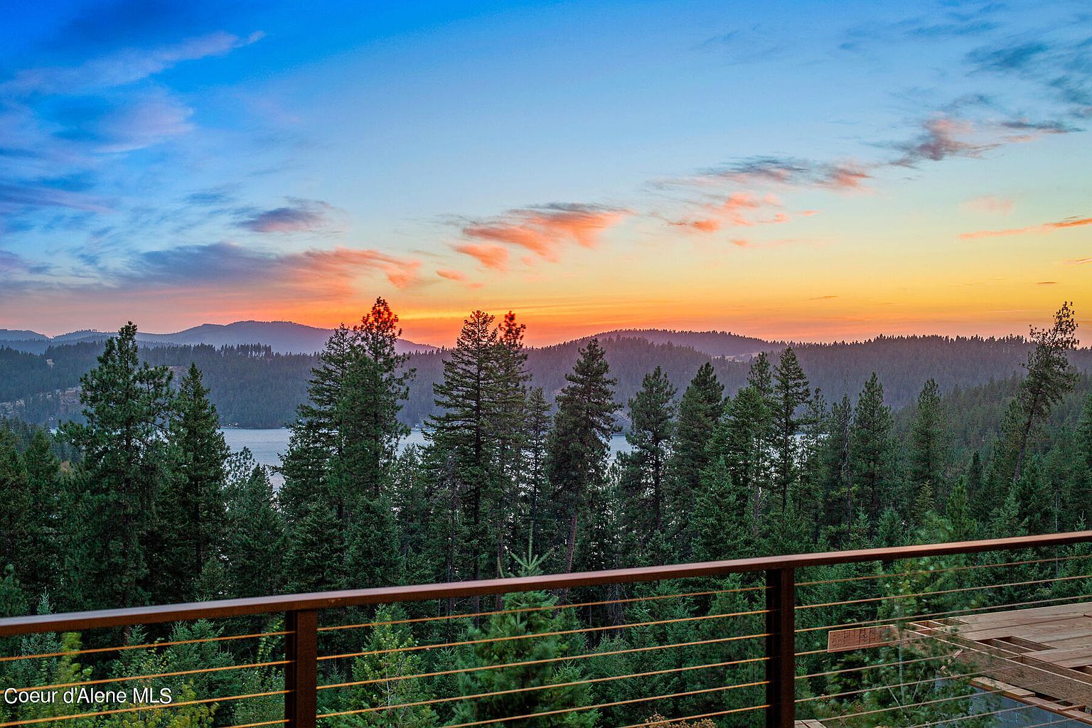 This image showcases a scenic view from a patio or balcony, featuring a lush forest landscape and a serene lake in the distance. The sky is painted with vibrant hues of orange and blue during sunset, creating a tranquil and picturesque setting. A wooden railing adds a touch of rustic charm to the foreground, enhancing the overall appeal of this outdoor living space.