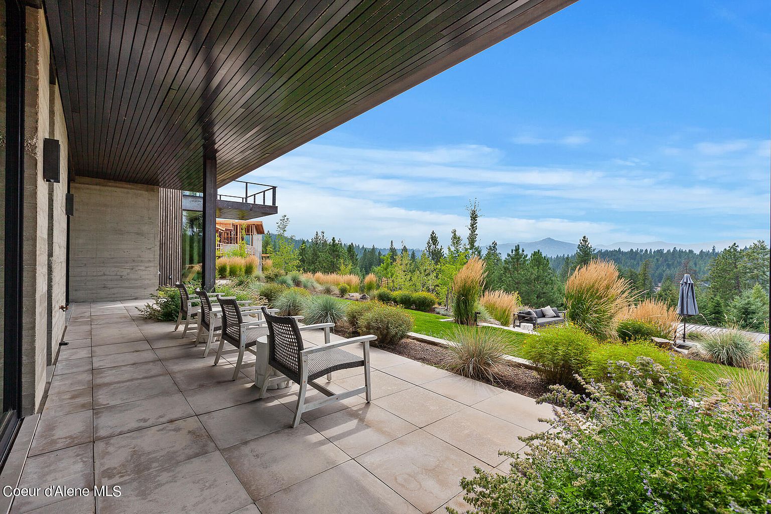 This image showcases a spacious patio or deck area with modern outdoor furniture, offering a serene view of a landscaped garden and distant mountains. The patio features large format tiles and a covered area with dark wood ceiling, creating a stylish and comfortable outdoor living space. The well-maintained garden adds to the property's appeal, making it an ideal spot for relaxation and entertainment.