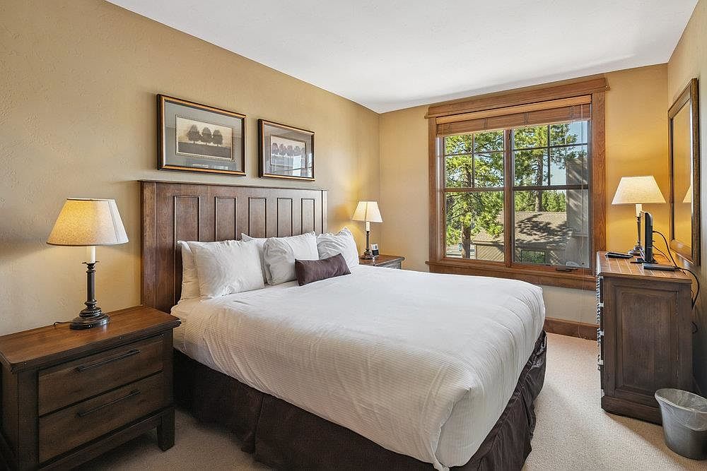 This is a cozy primary bedroom featuring a king-sized bed with a white duvet and brown accents. The room is decorated in warm, neutral tones with wooden furniture, including a nightstand with a lamp and a dresser. A large window provides natural light and a view of the outdoors, creating a relaxing and inviting atmosphere.