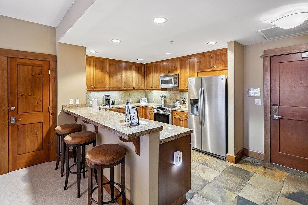 This is an interior shot of a kitchen featuring wooden cabinets, stainless steel appliances, and a granite countertop breakfast bar with three stools. The kitchen has a warm and inviting feel, with recessed lighting illuminating the space. The flooring is a multi-toned tile, adding visual interest to the room.