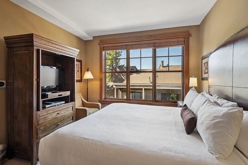 This is a cozy primary bedroom featuring a large bed with white linens and decorative pillows. A wooden armoire houses a television, and a window provides natural light and a view of the outdoors. The room has a warm, inviting atmosphere with neutral wall colors and comfortable furnishings.