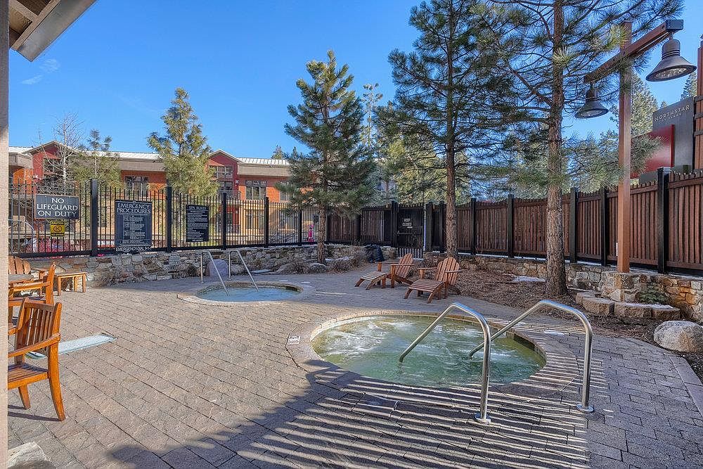 This image showcases an outdoor spa area with two hot tubs, surrounded by a brick patio and wooden fencing. Mature trees provide a natural backdrop, and lounge chairs are arranged for relaxation. Signage indicates pool rules and emergency procedures, suggesting a community amenity.