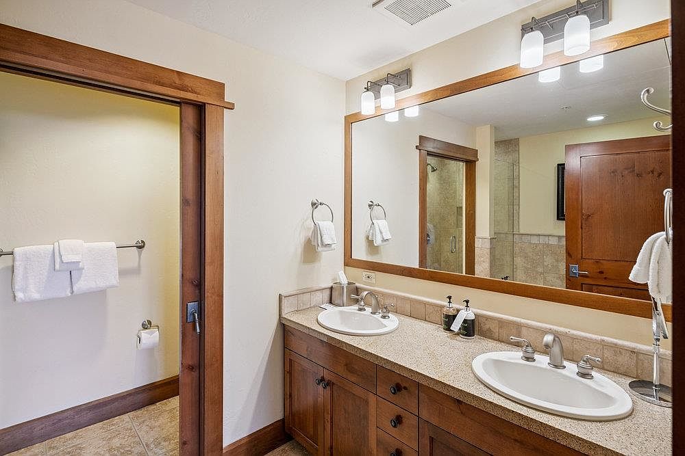 This is a well-lit primary bathroom featuring a double vanity with a light-colored countertop and dark wood cabinetry. A large mirror with integrated lighting hangs above the vanity, reflecting a glimpse of the shower and another door. The bathroom has a warm and inviting feel, with neutral wall colors and tiled flooring.