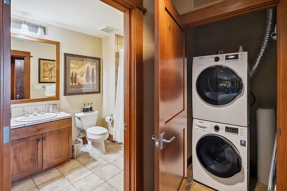 This image showcases a guest bathroom with a warm and inviting atmosphere. The bathroom features a wooden vanity with a white countertop and sink, a toilet, and a shower with a neutral-toned tile surround. Adjacent to the bathroom is a laundry area with stacked washer and dryer units, creating a convenient and space-saving design.