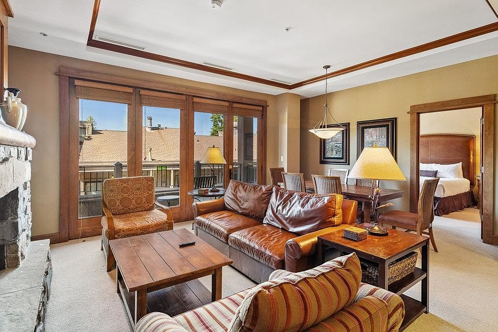 This inviting living room features a comfortable leather sofa, a patterned armchair, and a striped sofa, creating a cozy and stylish atmosphere. Natural light floods the space through large windows, highlighting the warm color palette and wooden accents. The room is well-appointed with a coffee table, side table, and a glimpse into the dining area and bedroom, suggesting a seamless flow within the home.
