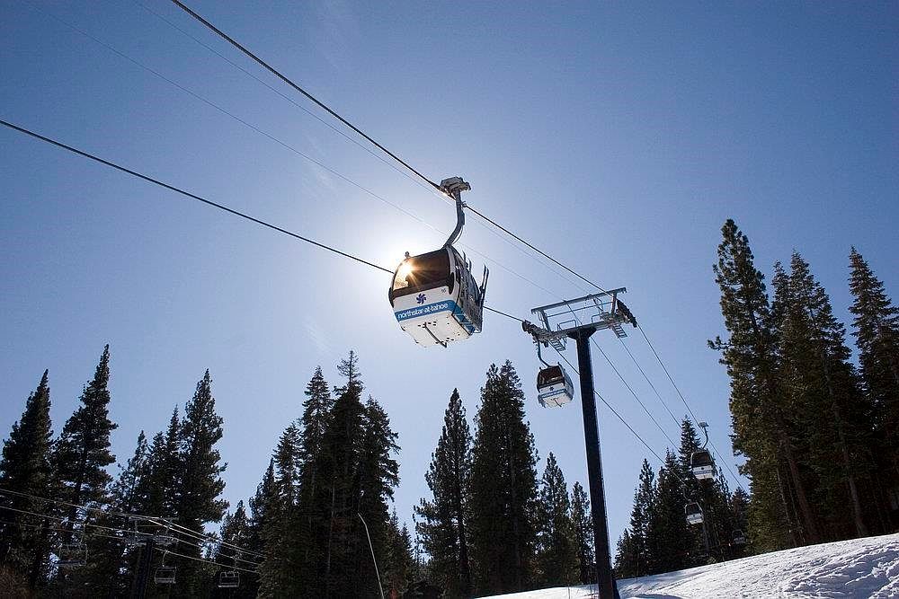 The image showcases a ski resort amenity, specifically a gondola lift system against a clear blue sky. Tall evergreen trees line the snowy slopes, creating a scenic winter landscape. The gondola cars are modern and appear well-maintained, suggesting a high-quality resort experience.