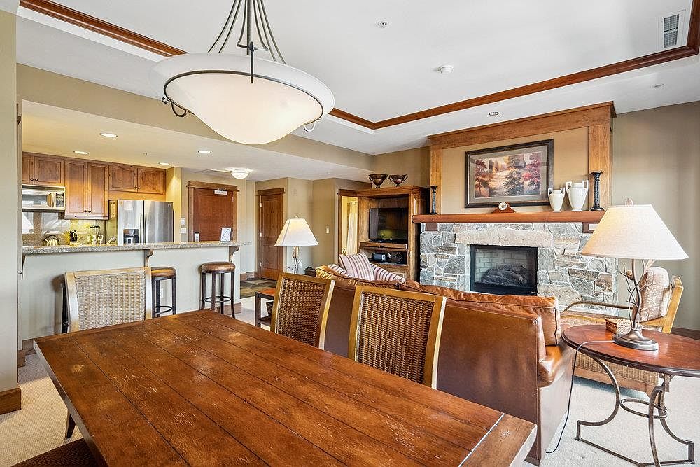This interior shot showcases a cozy living and dining area. The space features a stone fireplace, comfortable seating, and a wooden dining table with wicker chairs. The room is well-lit with a combination of natural light and stylish lamps, creating a warm and inviting atmosphere.