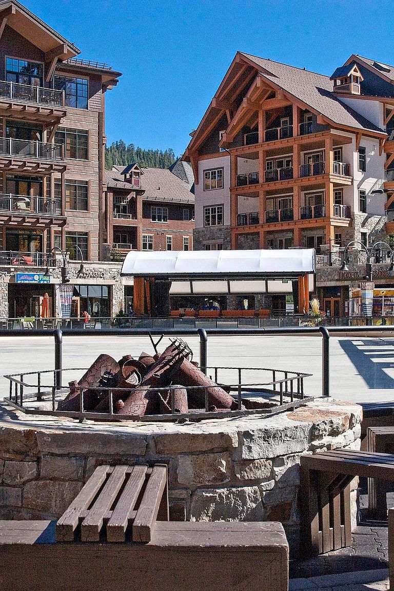 This image showcases the front view of a multi-story building complex with a rustic, mountain-lodge architectural style. The buildings feature a mix of wood and stone elements, with multiple balconies and large windows. In the foreground, there is a stone fire pit with metal accents, adding a cozy and inviting touch to the outdoor space.