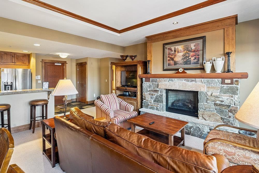 This living room features a stone fireplace with a wooden mantel, a brown leather sofa, and a striped armchair. A wooden coffee table sits in front of the sofa, and a television is built into a wooden cabinet. The room has a warm and inviting atmosphere with neutral wall colors and wooden trim.