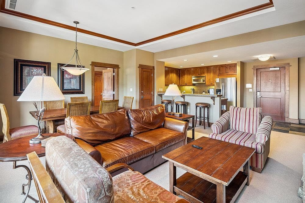 This is an interior shot of a living room that flows into a kitchen and dining area. The living room features a brown leather sofa, a patterned armchair, and a wooden coffee table. The kitchen has wooden cabinets and a breakfast bar with stools, while the dining area includes a wooden table with wicker chairs. The overall impression is warm and inviting, with a rustic yet comfortable style.