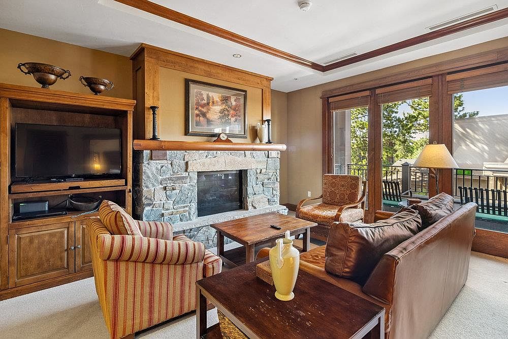 This is an interior shot of a living room featuring a stone fireplace, a built-in entertainment center, and comfortable seating. The room has a warm and inviting atmosphere with natural light coming through the sliding glass doors that lead to a balcony. The decor is traditional with wooden accents and earth-tone colors.