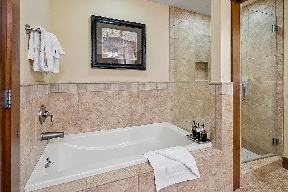 This is a well-appointed primary bathroom featuring a soaking tub with tile surround and a glass-enclosed shower. The neutral color palette and tile work create a spa-like atmosphere. A framed picture hangs above the tub, and toiletries are neatly arranged on a tray.