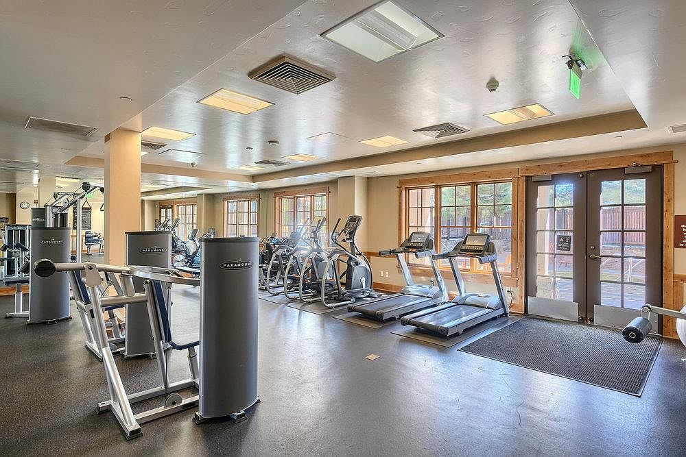 This is an interior shot of a well-equipped home gym. The room features several pieces of exercise equipment, including treadmills, elliptical machines, and weight training stations. Large windows provide natural light, and the overall impression is clean and functional, perfect for a health-conscious buyer.