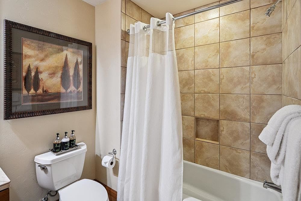 This is a bathroom featuring a toilet with toiletries on top, a framed picture on the wall, and a bathtub with a white shower curtain. The shower has tiled walls and a built-in niche. The overall impression is clean and functional, suitable for a guest bathroom.