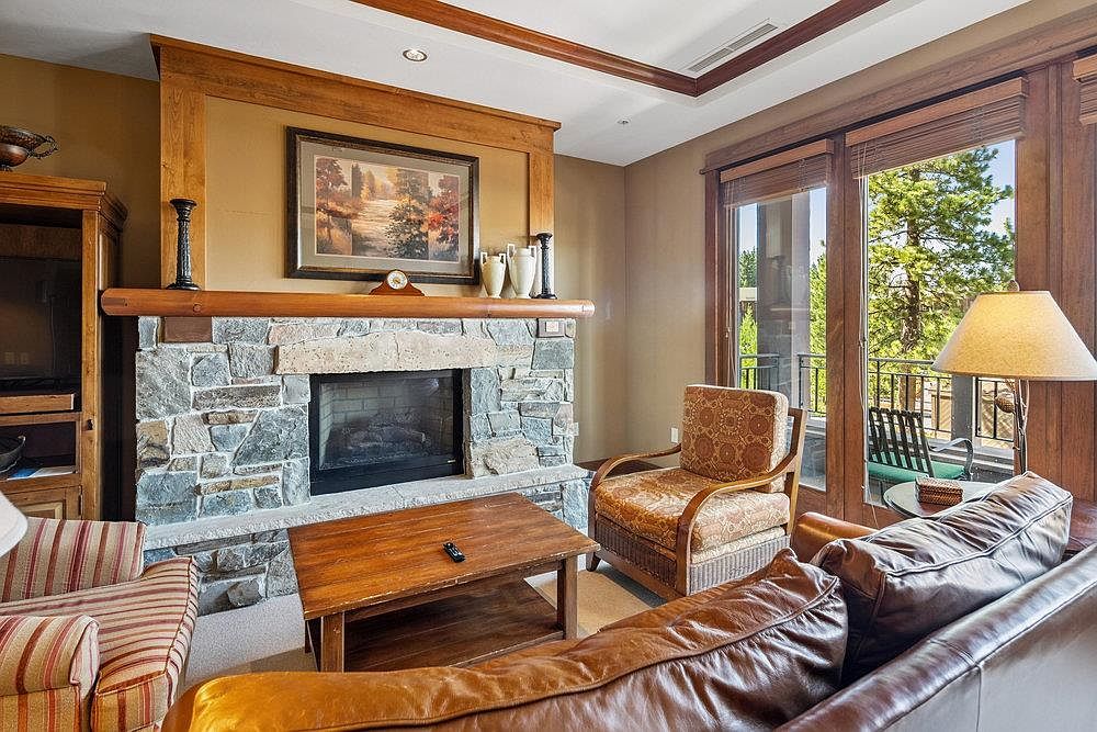 This is an interior shot of a cozy living room featuring a stone fireplace as a focal point, complemented by wooden accents and a painting above the mantel. Comfortable seating arrangements, including a leather sofa and patterned armchair, invite relaxation, while natural light streams in through the sliding glass doors leading to a balcony. The room exudes a warm and inviting atmosphere, perfect for a family home.