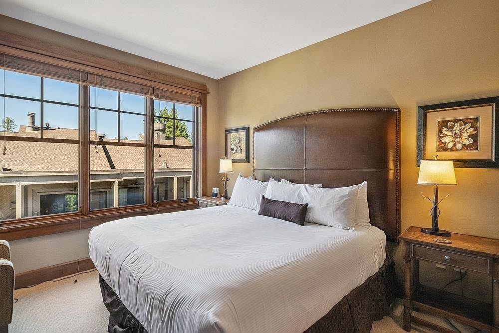 This is a cozy bedroom featuring a large bed with white linens and a brown headboard. A wooden nightstand with a lamp sits beside the bed, and a window provides natural light. The room has a warm and inviting atmosphere, perfect for a guest room.