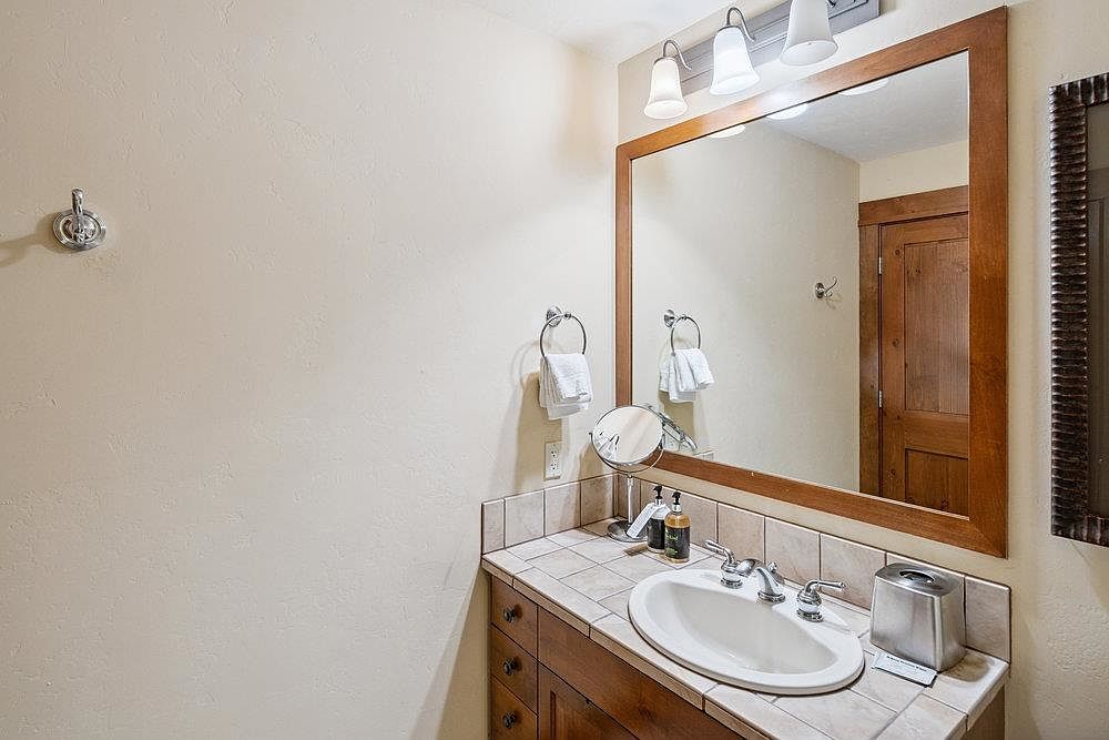 This is a well-lit bathroom featuring a wooden-framed mirror above a vanity with a light-colored countertop and wooden cabinets. A white oval sink is set into the countertop, complemented by chrome fixtures. The walls are painted in a neutral tone, and a towel rack with towels is visible, creating a clean and functional space.