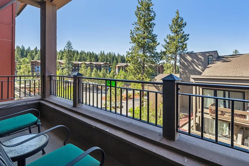 This image showcases a balcony with a black metal railing, offering a view of the surrounding trees and buildings. The balcony features outdoor seating with green cushions and a small glass-topped table. The overall impression is one of a relaxing outdoor space with a pleasant view.