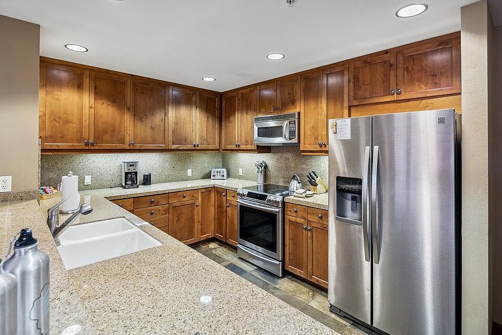 This is a well-lit kitchen featuring wooden cabinetry and granite countertops. Stainless steel appliances, including a refrigerator, microwave, and oven, are present. The kitchen has a warm and inviting feel, with ample counter space and storage.