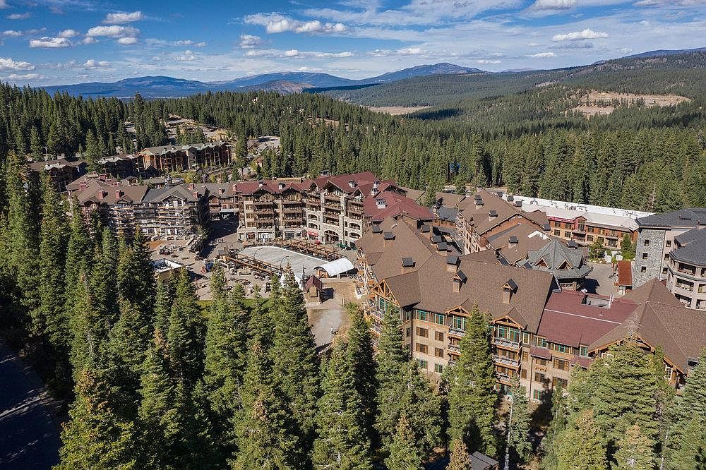 This aerial shot showcases a luxurious mountain resort nestled amidst a dense evergreen forest. The buildings feature a mix of stone and wood construction with steeply pitched roofs, suggesting a cozy, alpine aesthetic. A central plaza or courtyard area is visible, possibly offering amenities or gathering spaces for residents and guests.
