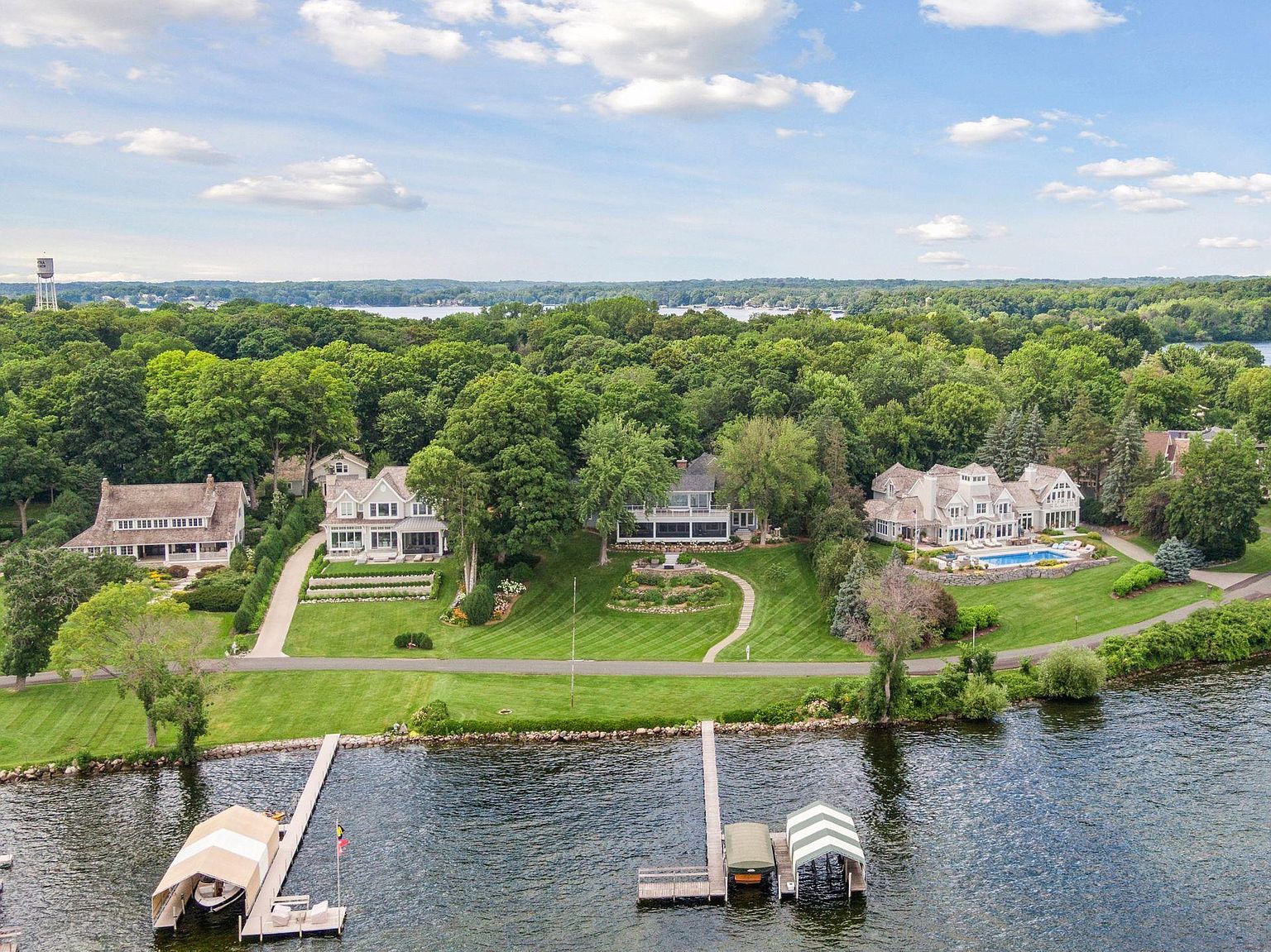 This aerial view showcases several luxurious waterfront homes with meticulously manicured lawns extending to the water's edge. Each property features unique architectural details and private docks, with lush green trees providing a serene backdrop. The scene conveys a sense of exclusivity and high-end living.