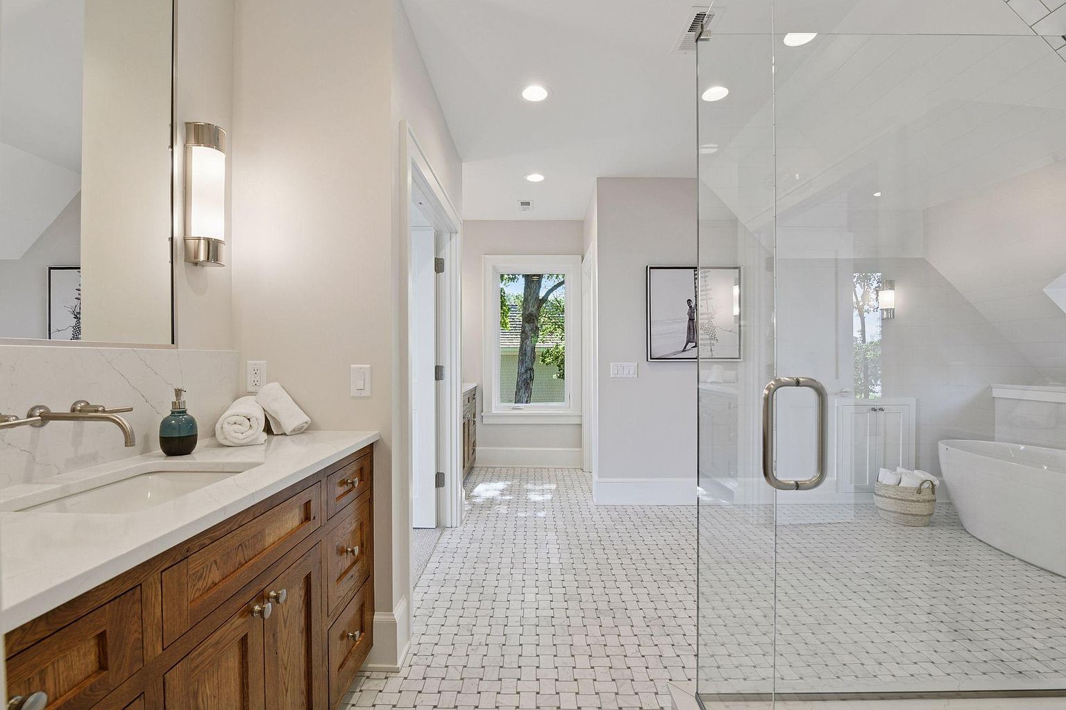 This is a bright and luxurious primary bathroom featuring a double vanity with a white countertop and wooden cabinets. The floor is covered in a mosaic tile pattern, leading to a glass-enclosed shower and a freestanding bathtub. The space is well-lit with recessed lighting and natural light from a window, creating a spa-like atmosphere.