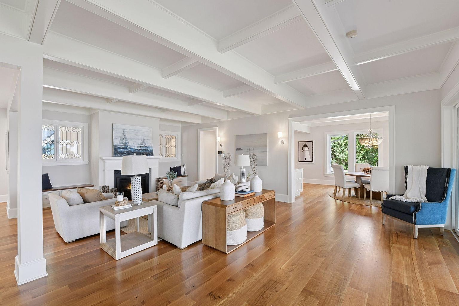This is an interior shot of a well-lit living room featuring hardwood floors, white coffered ceilings, and neutral-toned furniture. The room includes a fireplace, a console table with decorative vases, and an open doorway leading to a dining area. The overall impression is one of spaciousness and refined elegance.