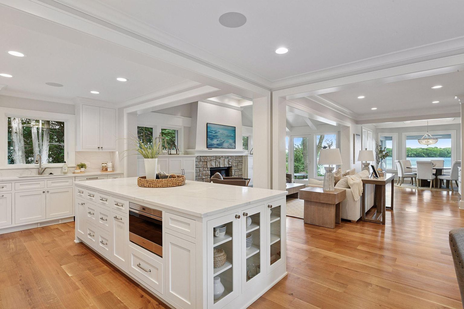 This is a bright and open kitchen that seamlessly flows into the living and dining areas. The kitchen features a large white island with a built-in oven and glass-front cabinets, complemented by hardwood flooring. The open-concept design enhances the sense of space and connectivity, making it ideal for entertaining and family living.