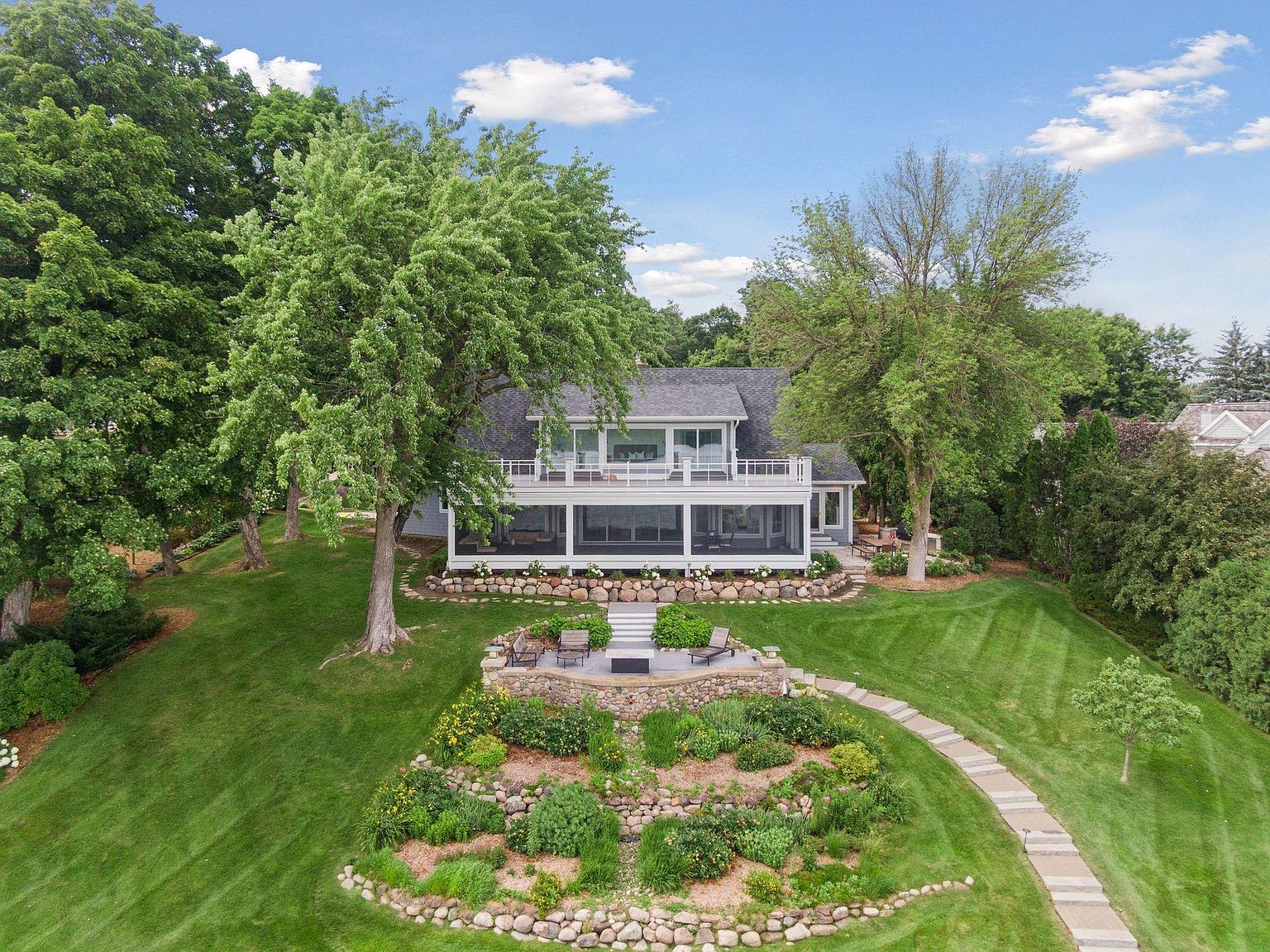 This aerial view showcases a beautiful two-story home with a large screened-in porch and an upper-level balcony. The property features meticulously maintained landscaping, including a stone-terraced garden and a winding stone pathway. Mature trees surround the house, providing shade and privacy, enhancing the overall appeal of this idyllic setting.