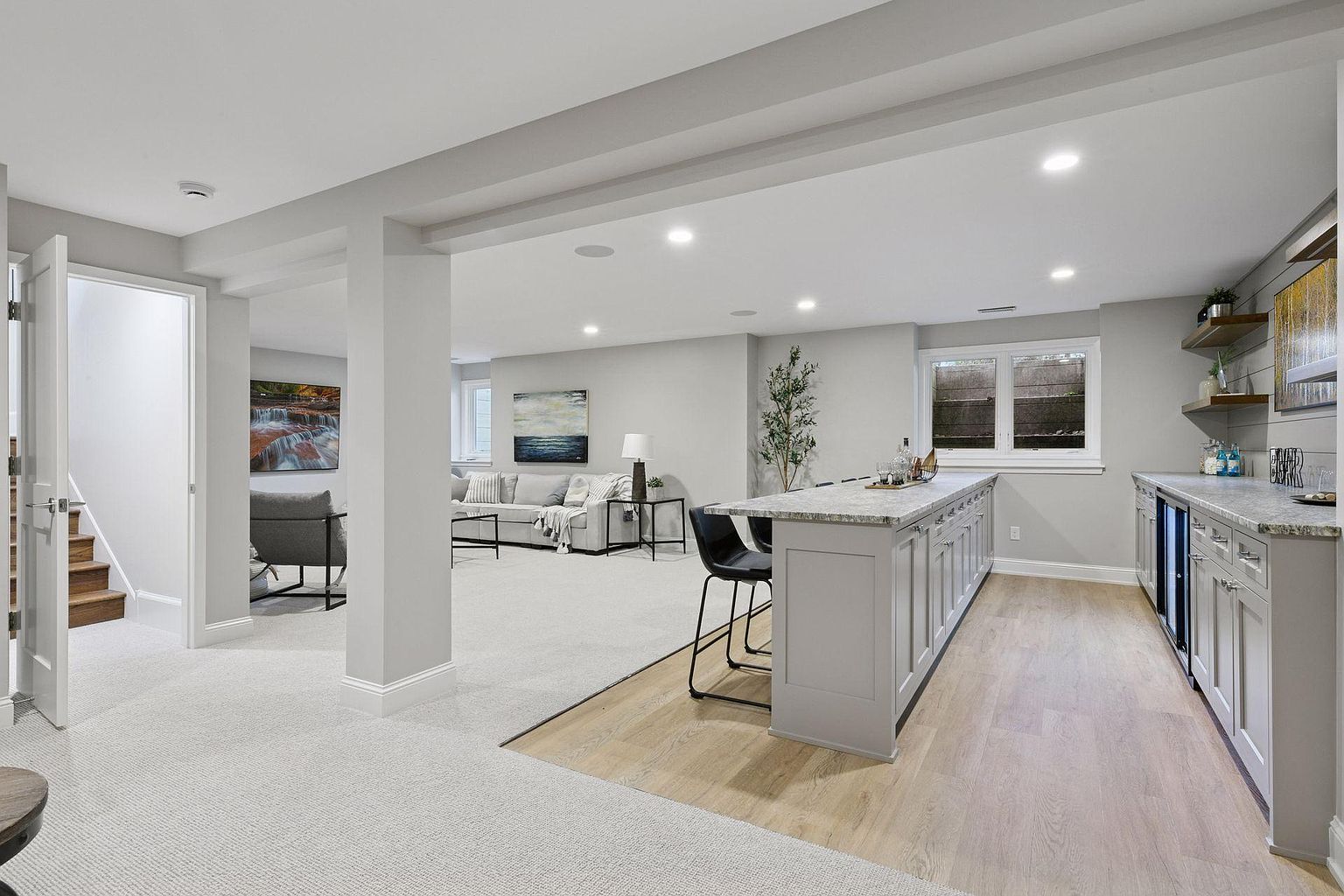 This is a well-lit basement featuring a living area with a gray sofa and artwork, a bar area with counter seating, and a beverage refrigerator. The flooring transitions from carpet in the living area to hardwood in the bar area, creating distinct zones within the open space. The overall impression is a modern and inviting finished basement.
