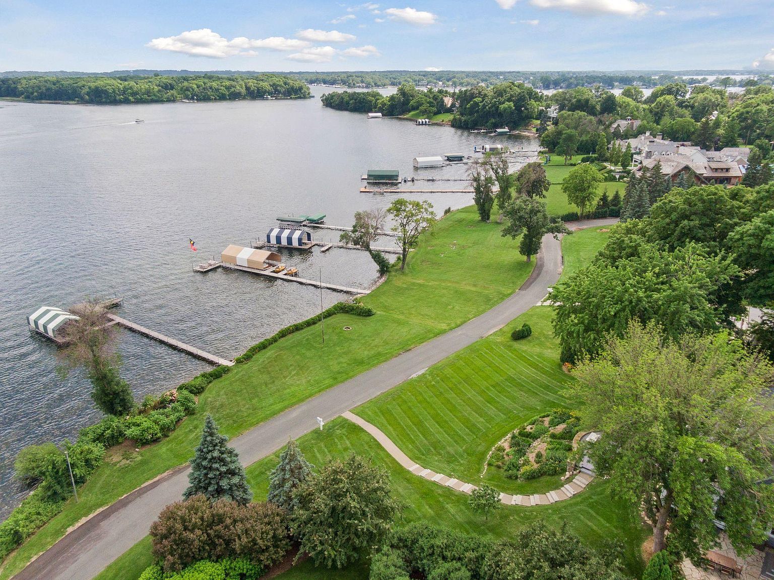 This aerial shot showcases a stunning waterfront property with lush green lawns, mature trees, and a private road leading to the main residence. Several docks and boat houses line the water's edge, offering direct access to the lake. The overall impression is one of luxury, privacy, and natural beauty.