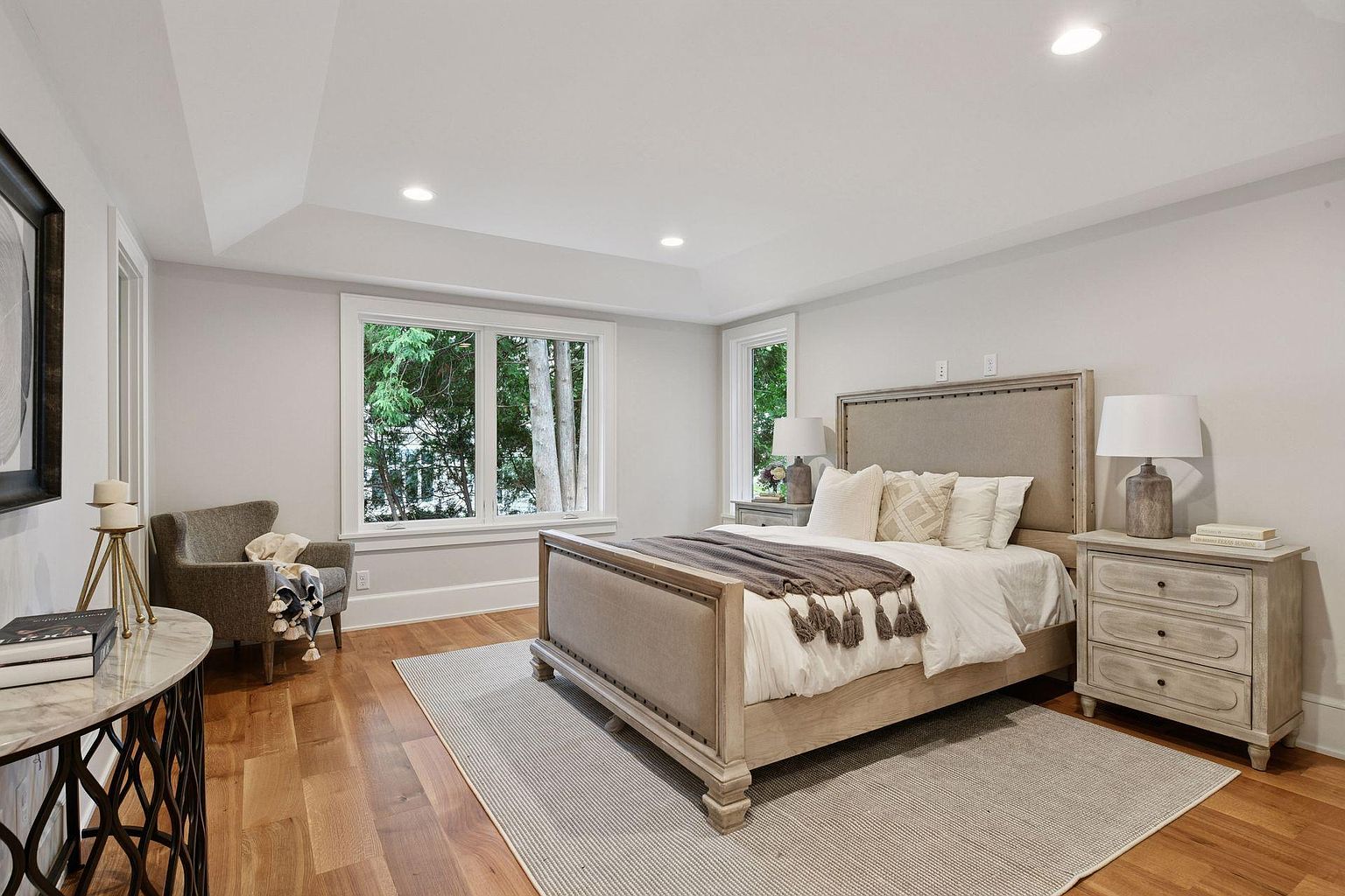 This is a well-lit primary bedroom featuring a neutral color palette. The room includes a large bed with a padded headboard, matching nightstands with lamps, and a comfortable armchair. A large window provides natural light and a view of the outdoors, enhancing the serene and inviting atmosphere.