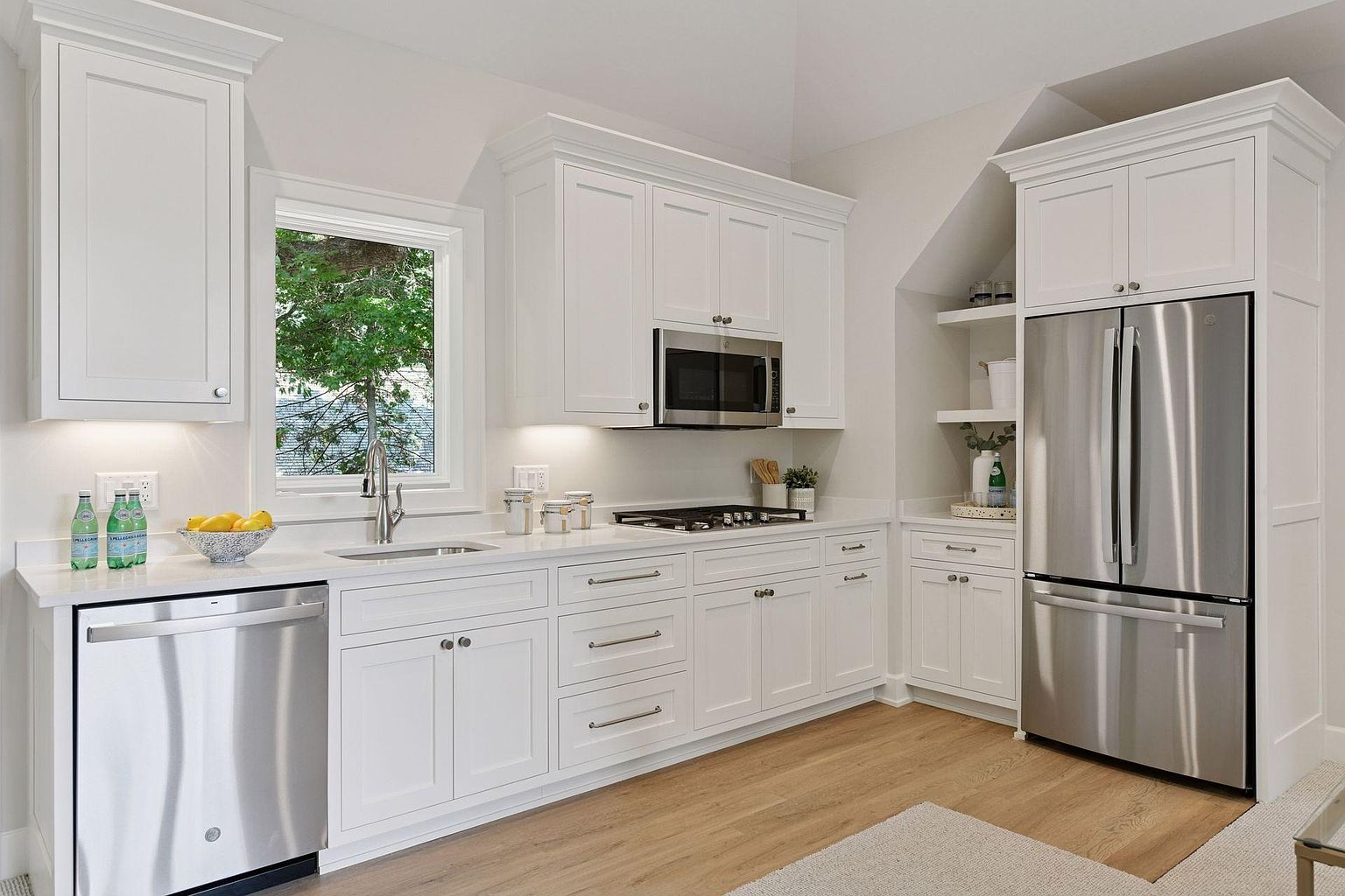 This is a bright, modern kitchen featuring white cabinetry, stainless steel appliances, and light wood flooring. The kitchen is well-lit, with natural light coming in from a window above the sink. The perspective is from a medium distance, capturing the entire kitchen layout and creating a welcoming and functional space.