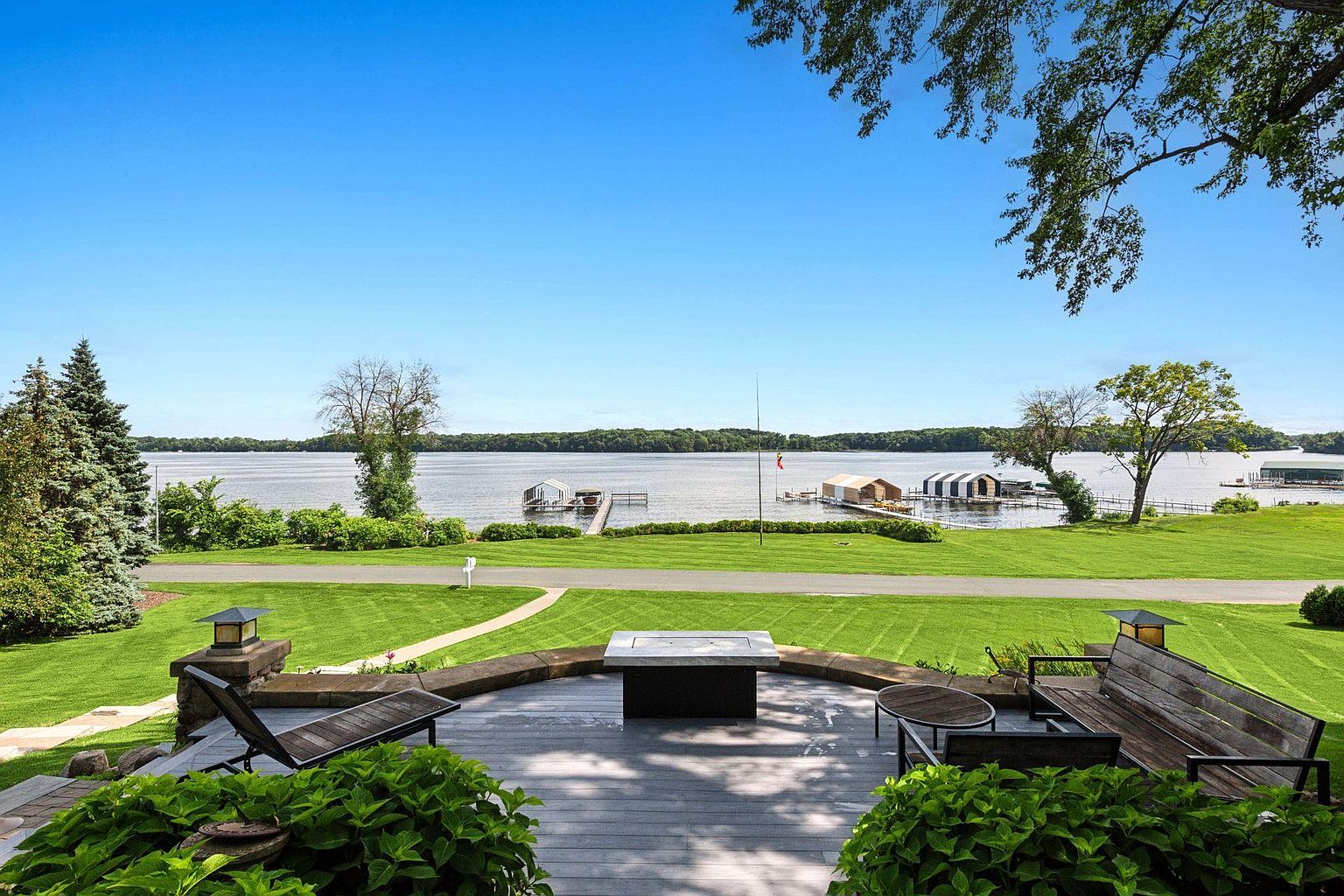 This image showcases a beautifully designed outdoor patio or deck area with a stunning lake view. The deck features comfortable seating arrangements, including a lounge chair and a wooden bench, centered around a modern fire pit. The well-manicured lawn extends towards the lake, enhancing the property's appeal and creating a serene outdoor living space.