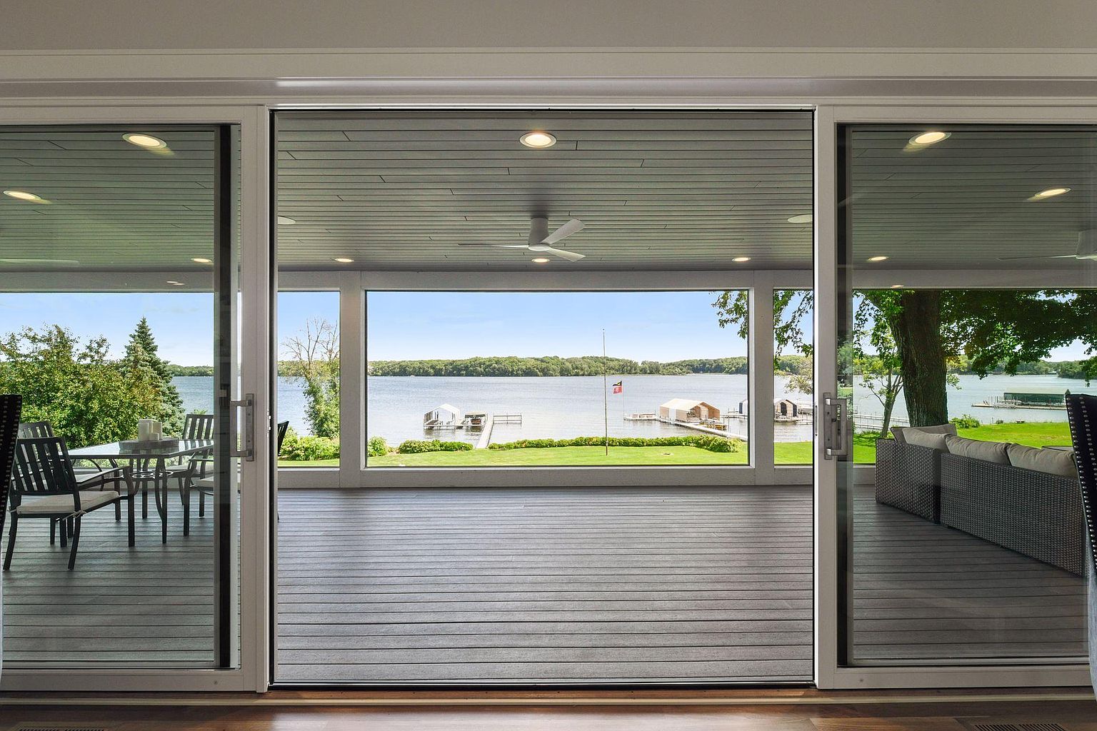 This image showcases a spacious outdoor patio or deck area, seamlessly connected to the interior through large sliding glass doors. The deck features modern, horizontally-laid decking material and offers stunning views of a lake with boat docks and lush greenery. The space is designed for relaxation and entertainment, with comfortable seating visible on one side and an outdoor dining area on the other.