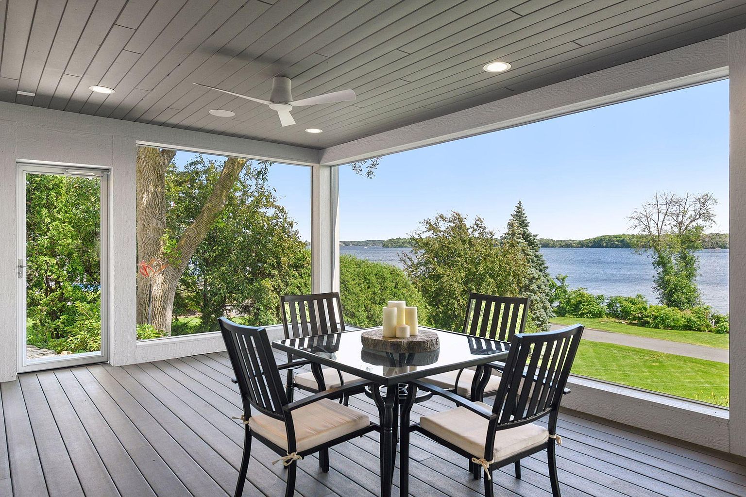 This image showcases a screened-in porch or patio area, furnished with a black dining table and four chairs, creating an inviting outdoor dining space. The space is framed by large windows offering scenic views of a lake and lush greenery, enhancing the appeal of the property. The ceiling features recessed lighting and a ceiling fan, adding to the comfort and functionality of the area.