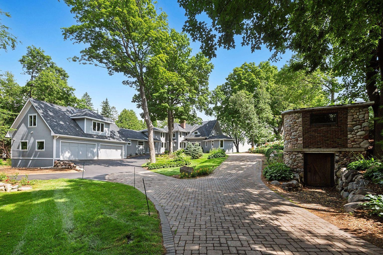 This image showcases the front exterior of a well-maintained home with a long, winding brick driveway leading up to the property. The house features a gray exterior with a multi-car garage and is surrounded by lush greenery, including mature trees and manicured lawns. A unique stone and brick structure adds character to the landscape, suggesting a blend of rustic charm and modern living.