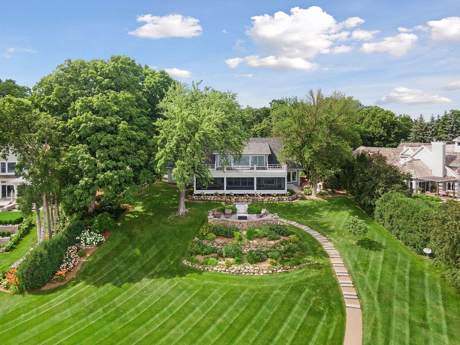 This aerial view showcases a beautifully landscaped property featuring a large house with a multi-level deck. The meticulously maintained lawn is adorned with garden beds and a stone pathway leading to the house. Mature trees surround the property, providing privacy and a lush, green backdrop.