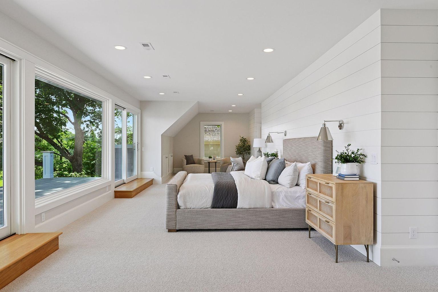This is a bright and spacious primary bedroom featuring a large bed with neutral linens and a gray accent throw. The room has a modern farmhouse aesthetic with white shiplap walls and a light wood nightstand. Large windows offer ample natural light and views of the surrounding greenery, enhancing the serene atmosphere.