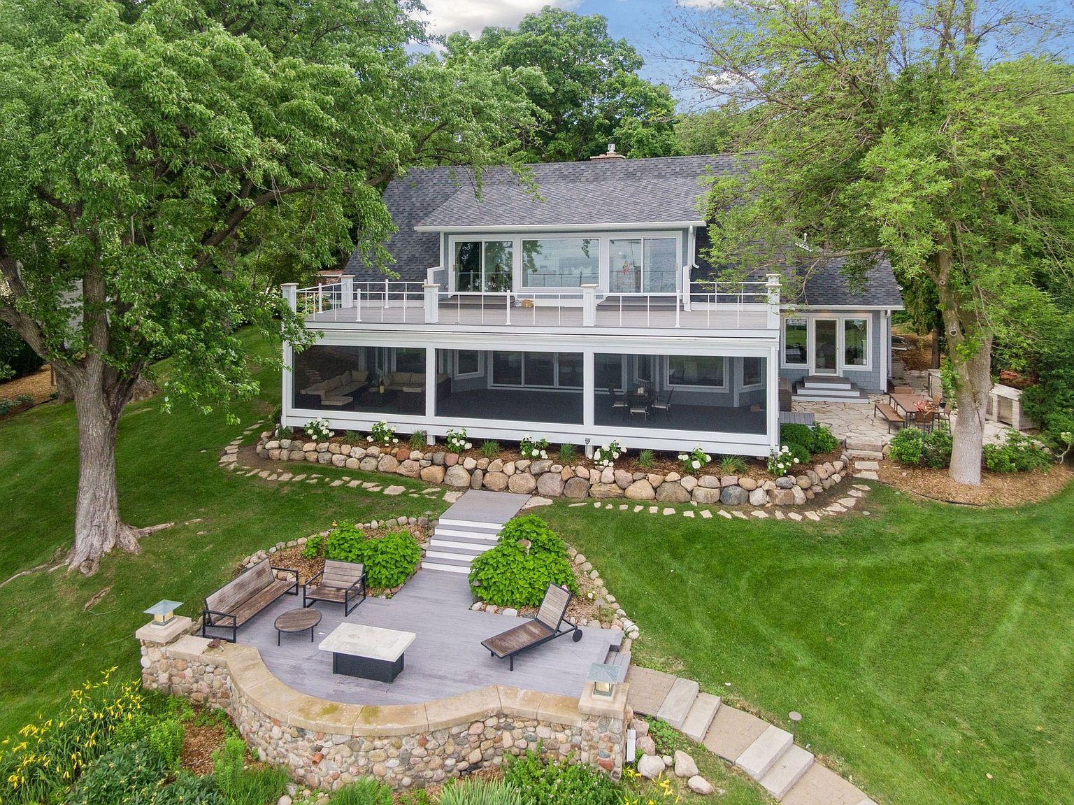 This is an aerial rear view of a two-story house with a large deck and patio area. The house features a gray roof, light blue siding, and large windows. The deck is spacious with white railings, and the patio below is screened in. A stone retaining wall and landscaping add to the property's appeal, creating an inviting outdoor living space.