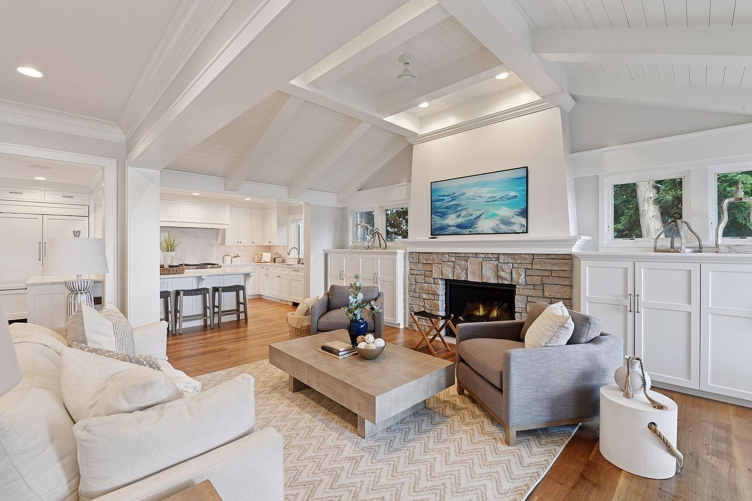 This is an interior shot of a living room that flows into a kitchen area. The living room features a stone fireplace with a mounted television, comfortable seating, and a neutral-toned rug. The kitchen has white cabinetry and a breakfast bar, creating an open and inviting space.