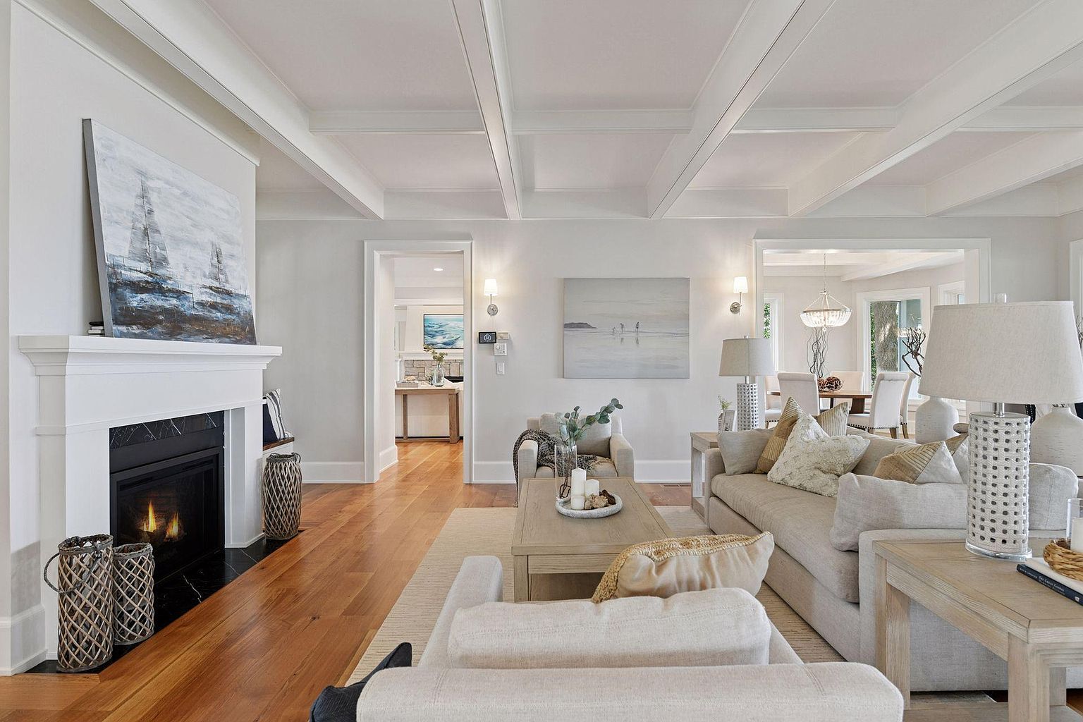 This is an interior shot of a living room featuring a fireplace with a painting above it, hardwood floors, and neutral-toned furniture. The room has a bright and airy feel, enhanced by the natural light and the white coffered ceiling. The open layout connects to other rooms, suggesting a spacious and inviting home.