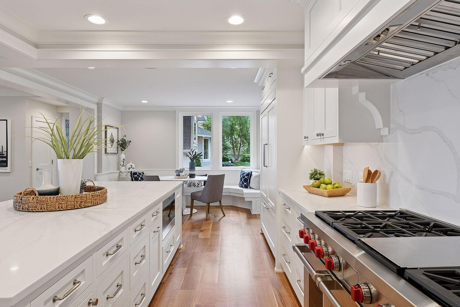 This is a bright and airy kitchen featuring white cabinetry, stainless steel appliances, and a large island with a white countertop. The kitchen has hardwood floors and a view into a dining area with a window overlooking the outdoors. The perspective is from the entrance of the kitchen, showcasing the layout and design.