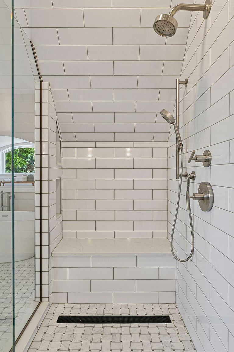 This is a well-lit primary bathroom shower featuring white subway tile walls and a mosaic tile floor. The shower includes a built-in bench, a rainfall showerhead, and a handheld shower fixture. The design is clean and modern, creating a spa-like atmosphere.