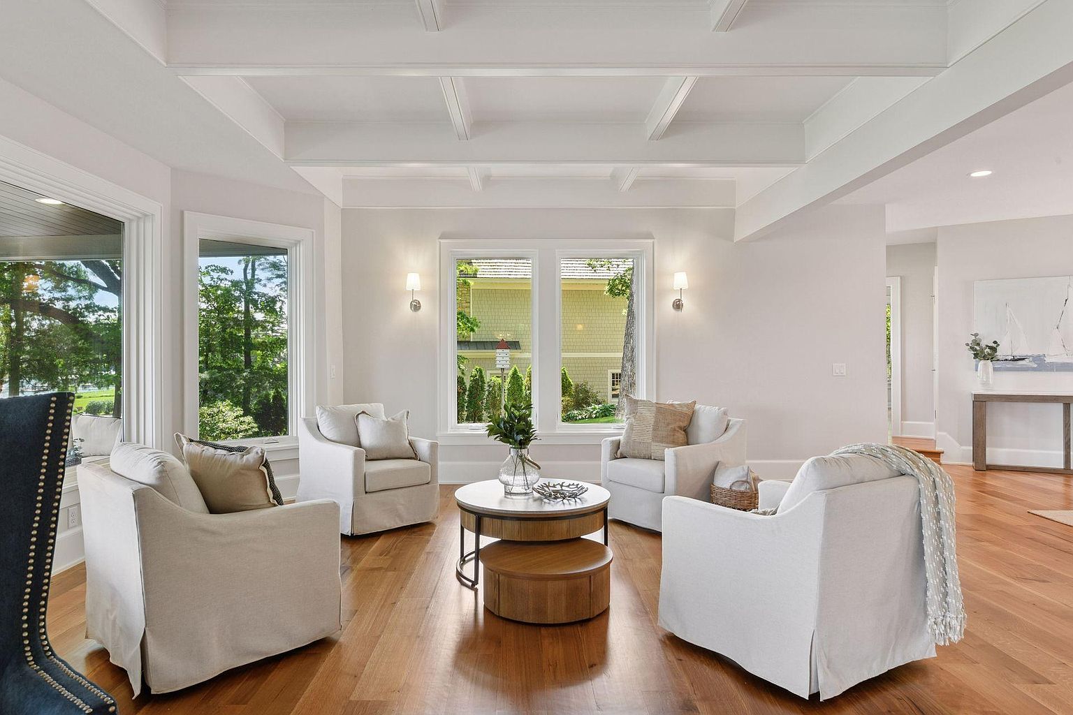 This is an inviting living room featuring four comfortable armchairs arranged around a two-tiered round coffee table. The room is bathed in natural light from the surrounding windows, offering views of the outdoors. The hardwood floors and coffered ceiling add a touch of elegance to the space, creating a cozy and sophisticated atmosphere.