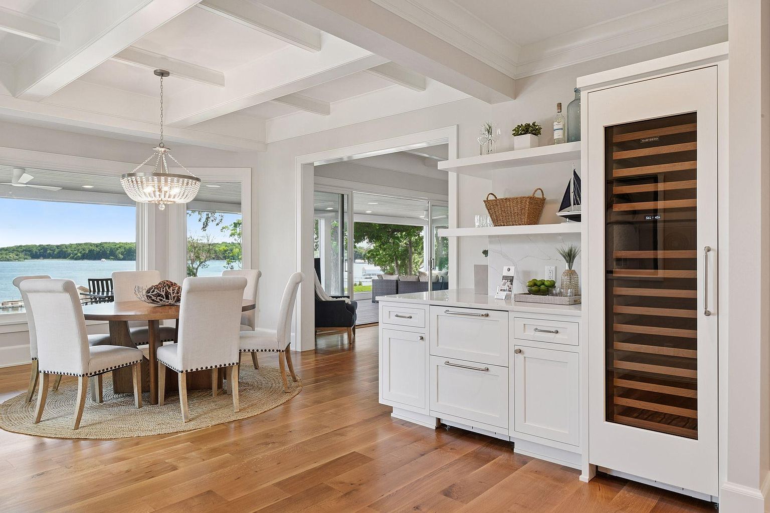 This is an interior shot of a dining room featuring a round wooden table surrounded by upholstered chairs with nailhead trim, set on a round jute rug. A chandelier hangs above the table, and a large window offers a view of the water. The room is bright and airy, with white walls and coffered ceilings, creating an elegant and inviting atmosphere.