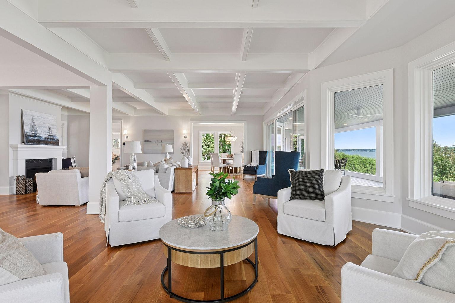 This is a bright and airy living room featuring hardwood floors, white walls, and a coffered ceiling. The room is furnished with several white armchairs, a blue accent chair, and a round coffee table with a glass vase and greenery. Large windows offer a view of the outdoors, creating a seamless connection between indoor and outdoor spaces.
