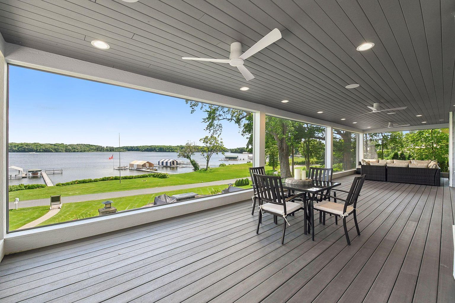 This image showcases a spacious outdoor patio or deck area with a stunning waterfront view. The deck features a dining set and comfortable lounge seating, perfect for relaxation and entertaining. The gray decking and ceiling create a modern and sophisticated look, while the large windows offer unobstructed views of the lake and surrounding landscape.