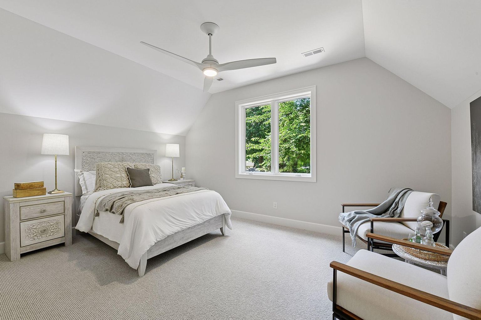 This is a bright and airy primary bedroom featuring a neutral color palette. The room includes a bed with decorative pillows and a throw blanket, a nightstand with a lamp, and a seating area with two chairs. A large window provides natural light and a view of the outdoors, enhancing the serene and inviting atmosphere.
