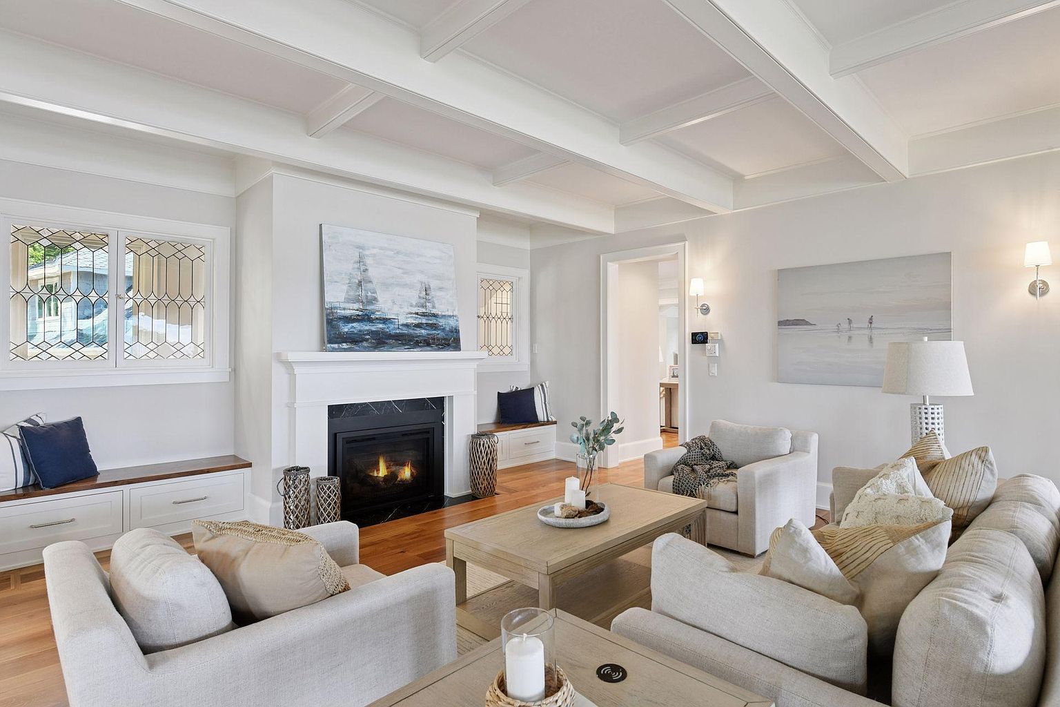This is a bright and airy living room featuring a coffered ceiling, a fireplace with a painting above it, and comfortable seating arrangements. The room is decorated in neutral tones with hardwood floors, creating a warm and inviting atmosphere. The perspective is from the front of the room, showcasing the fireplace and seating area.